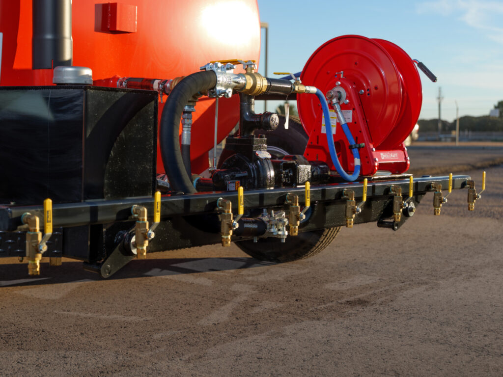 A spray bar with several yellow nozzles on the back of a black trailer with a red tank.